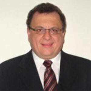 A imagem mostra o retrato de um homem de meia-idade posando para uma foto formal.Ele está de frente para a câmera, com uma expressão leve e sorridente. Tem cabelos curtos e escuros, com um pouco de volume nas laterais. Usa óculos de armação fina, que deixam os olhos bem visíveis. A roupa é formal: ele veste um terno escuro, camisa branca e uma gravata com listras diagonais em tons de vermelho e preto. O fundo da foto é liso e claro, sem objetos ou cenário ao redor, o que faz com que toda a atenção fique no rosto e na vestimenta da pessoa. A imagem lembra uma foto institucional ou profissional, como aquelas usadas em perfis de trabalho, currículos ou sites oficiais.