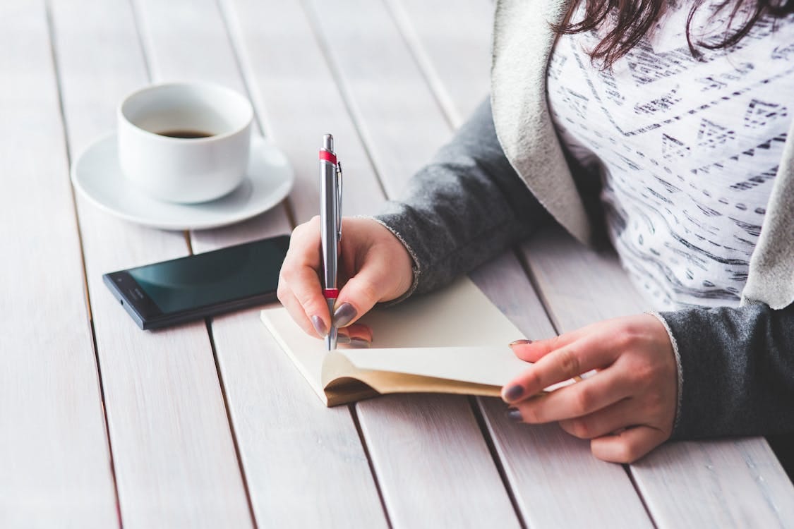 Free A woman writes in a journal next to a cup of coffee and a smartphone on a wooden table. Stock Photo