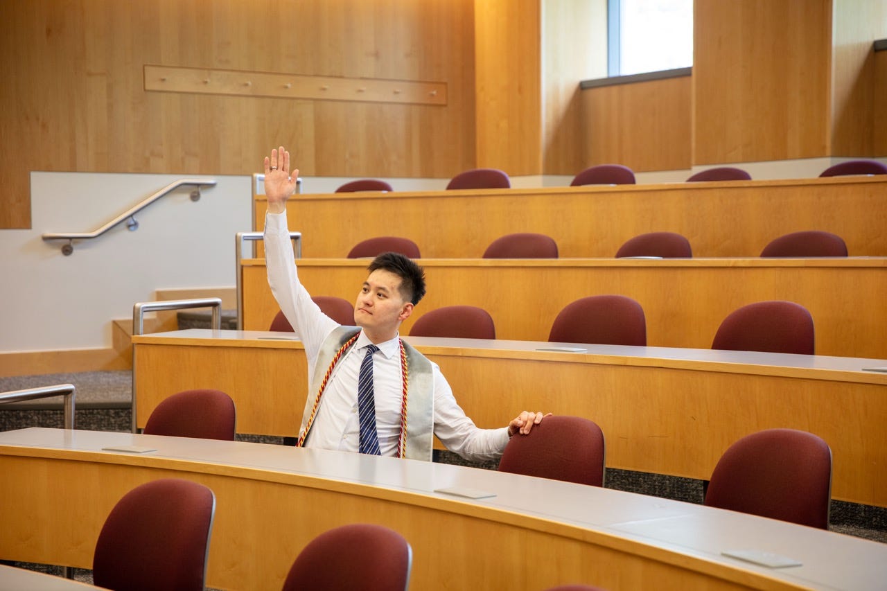 Junru wearing graduation cords and an LGO stole raises his hand in an empty Sloan classroom Junru wearing graduation cords and an LGO stole raises his hand in an empty Sloan classroom