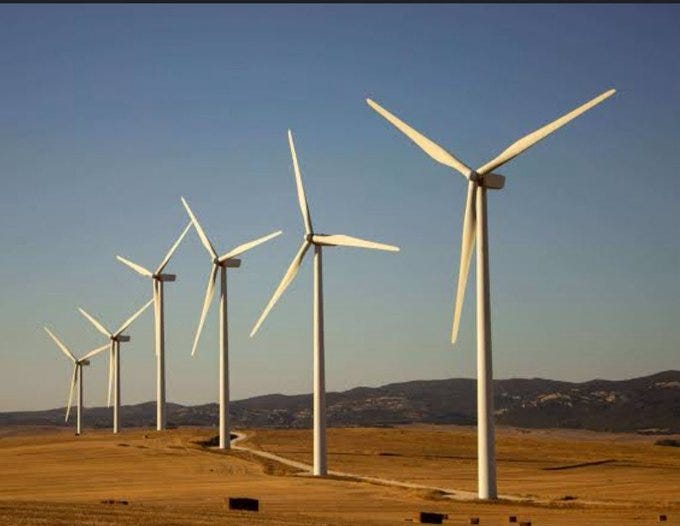 Multiple white wind turbines with three blades each stand in a row on a dry grassy field under a clear blue sky with distant brown hills and a dirt path running alongside them