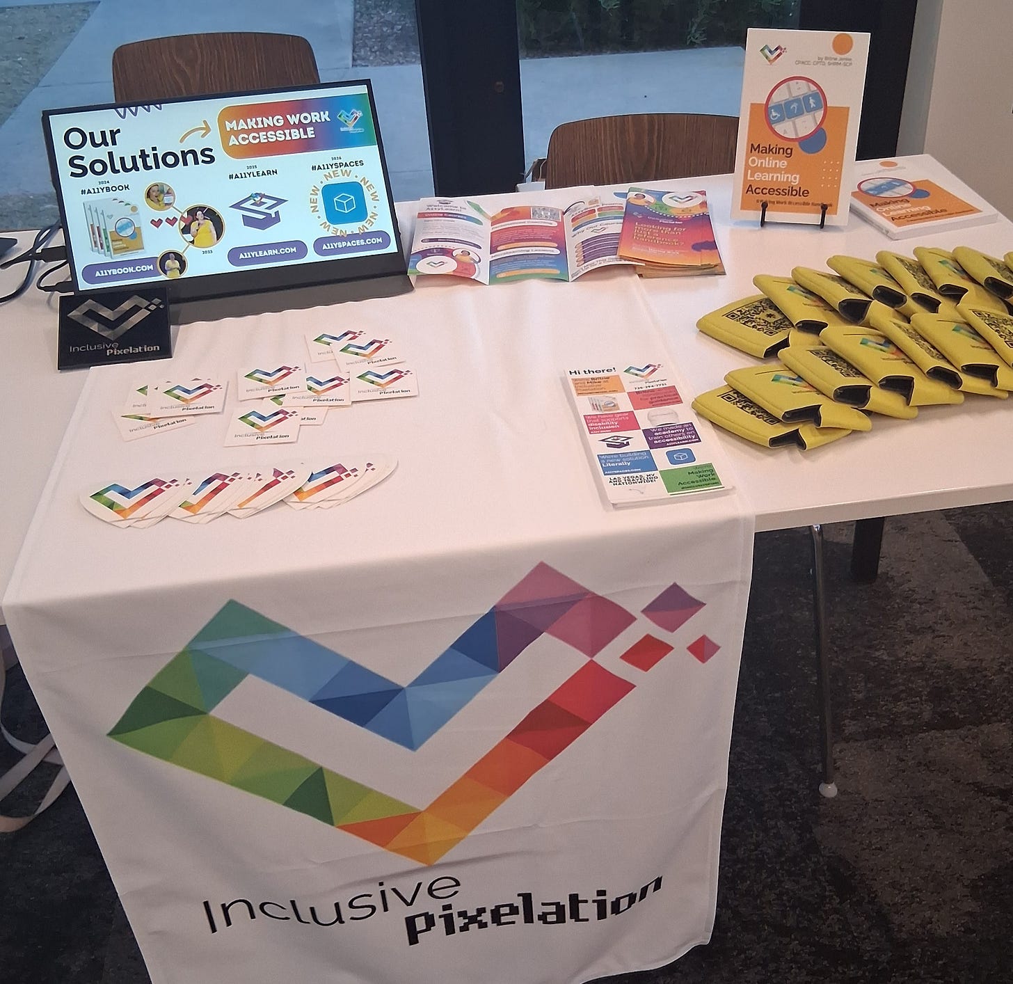 A small table with a white drape showing the Inclusive Pixelation rainbow heart logo. The table is covered in swag: books, brochures, can cozies, stickers, flyers, business cards, and a monitor displaying our services.