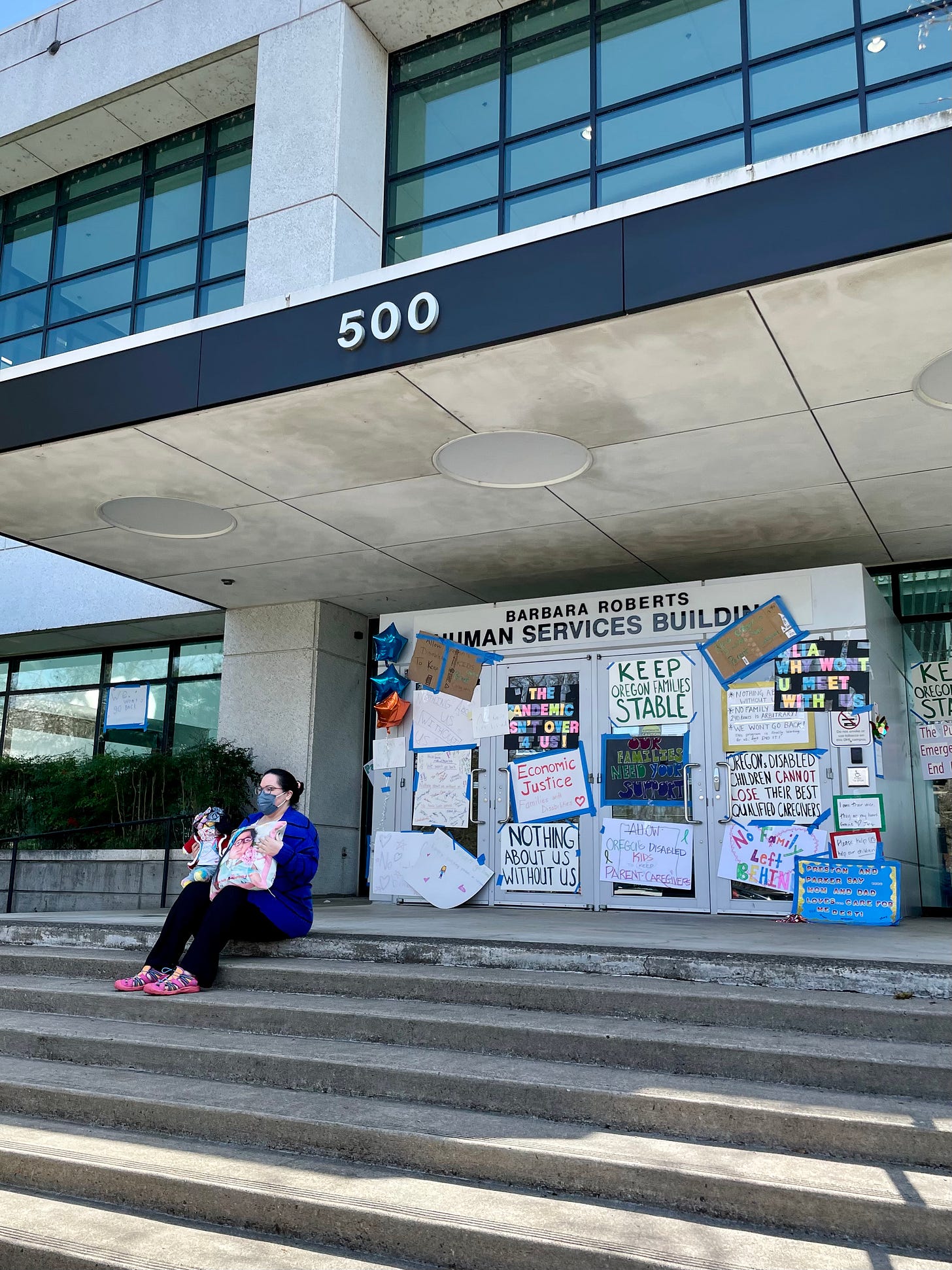 A woman sits on the steps in front of a large government building. Multicolored signs paper the door behind her.