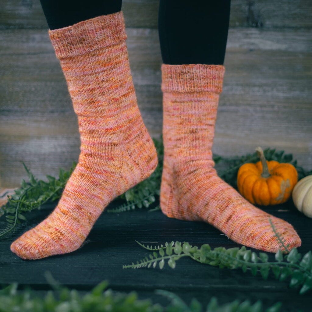 A pair of hand-knit socks in shades of peach and yellow stands against a rustic wooden background, flanked by green foliage and a small orange pumpkin, evoking a cozy, autumnal feel. A pair of hand-knit socks in shades of peach and yellow stands against a rustic wooden background, flanked by green foliage and a small orange pumpkin, evoking a cozy, autumnal feel.