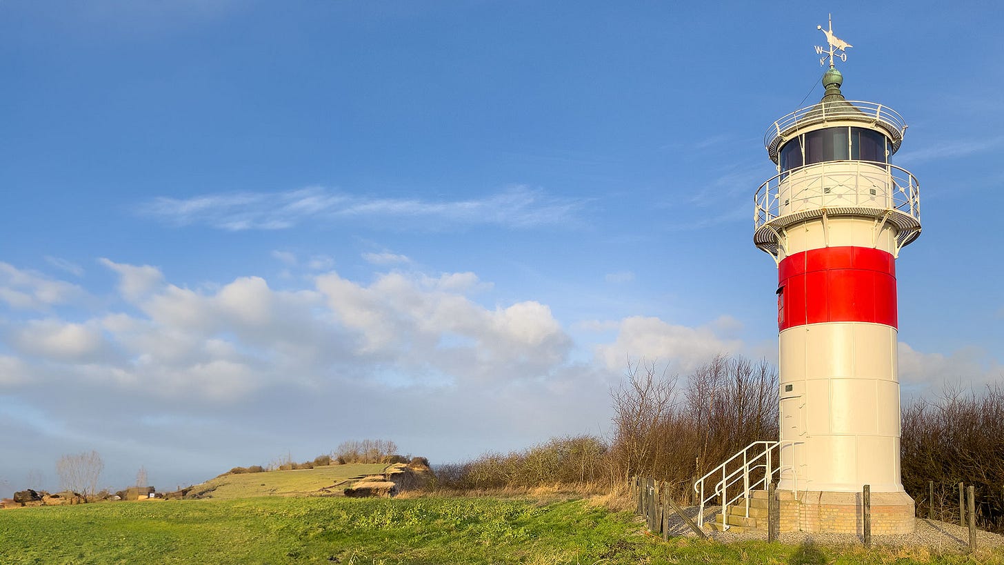 Ein kleiner weißer Leuchtturm mit einem breiten roten Streifen vor blauem Himmel und einer Wiese mit laublosen Bäumen und Sträuchern.