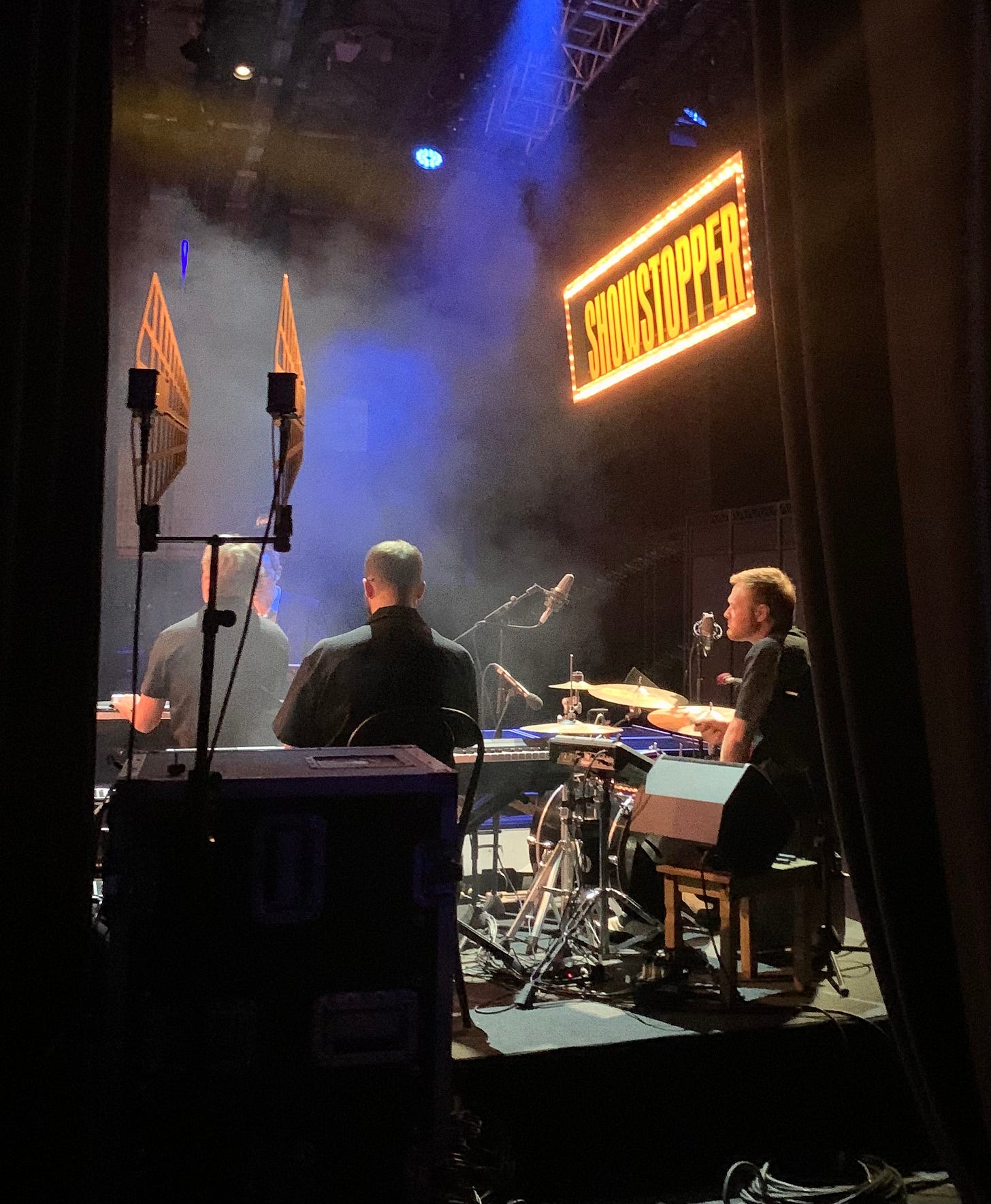 The backs of three men on stage, seen through the stage left wing. All are wearing black shirts. There is a yellow sign at the top of the stage saying "Showstopper"