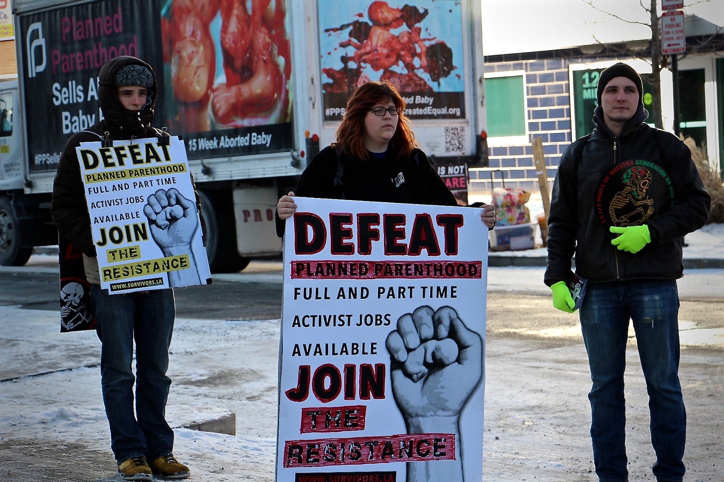 A photograph of anti-abortion activist Lauren Handy, who stands in the center of the image holding a protest sign, flanked on either side by two male protesters. A photograph of anti-abortion activist Lauren Handy, who stands in the center of the image holding a protest sign, flanked on either side by two male protesters.