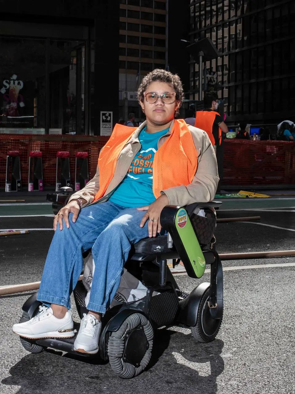 A photo of a disabled woman in her powerchair. She is brightly lit outside on a street, wearing an orange vest, teal shirt, jeans, and white shoes.