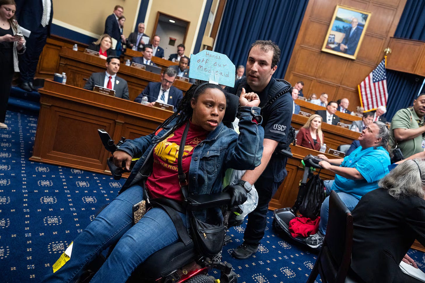 A protestor in a wheelchair is arrested in the committee chamber. Her fist raised in defiance. 