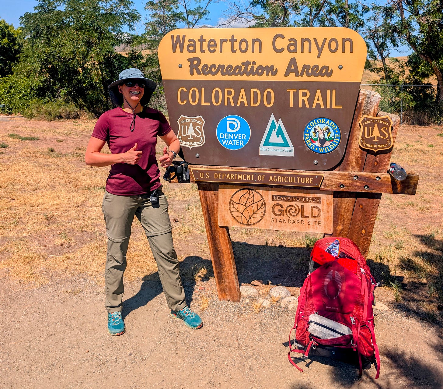 Thru-hiker smiling with trekking poles at the Waterton Canyon Colorado Trailhead sign, red Osprey pack propped against the post in the summer sun.