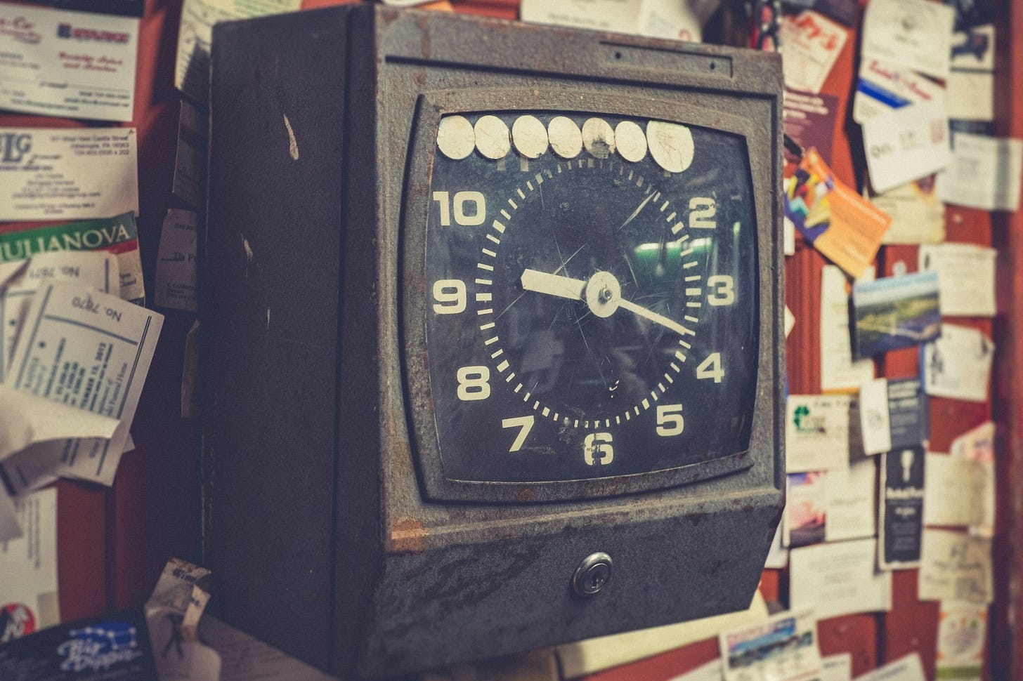 An old industrial punch clock with a black face and white numbers, mounted against a wall cluttered with papers and cards.