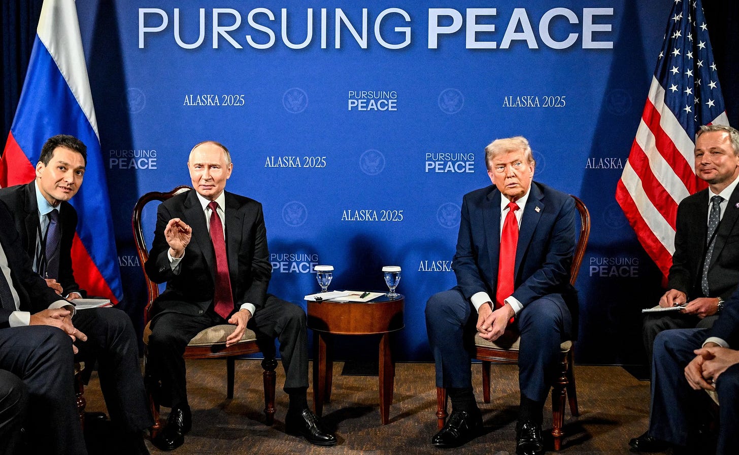 Putin and Trump sitting on two chairs, with a background behind them reading "pursuing peace", together with translators and other diplomats.