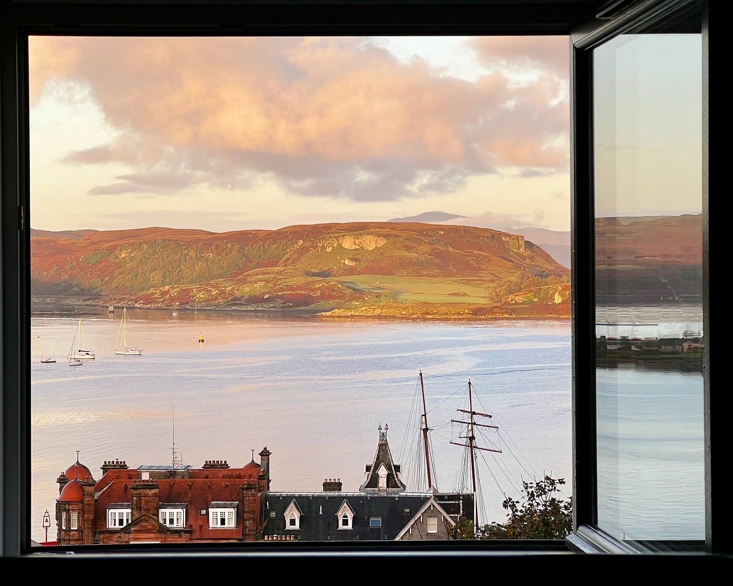 A black window frames a pink-blue calm sea with boats on it, with brown-green hills in the background and a pink cloudy sky - in the foreground are two buildings, red and black, and a spire and masts.