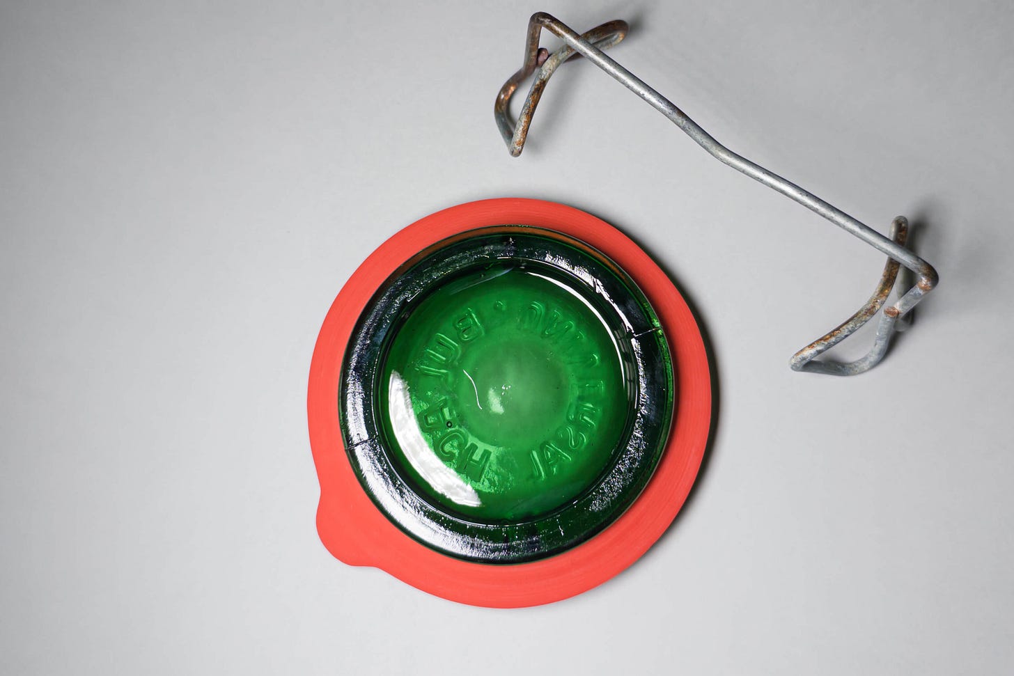 Top view of a green Buelach jar lid with an orange rubber ring seated around the rim; the separate metal swing clamp rests next to it, ready to lock things down old-school style. Top view of a green Buelach jar lid with an orange rubber ring seated around the rim; the separate metal swing clamp rests next to it, ready to lock things down old-school style.