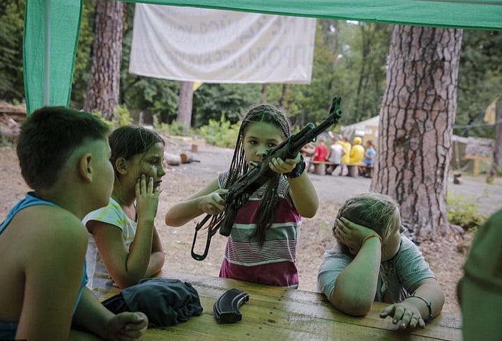 Pictures show young children being taught how to use rifles by Azov members with Swastika tattoos.