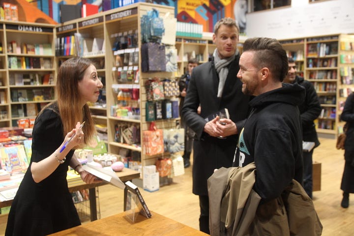 Author Isabelle Kenyon chats to two writers with a book and pen in her hand in a colourful bookshop (Blackwell's); In the second image author Isabelle hugs a reader with book 'The Dark Within Them' in her hand, in background the bookshop is selling cards on a portrait colourful rack.