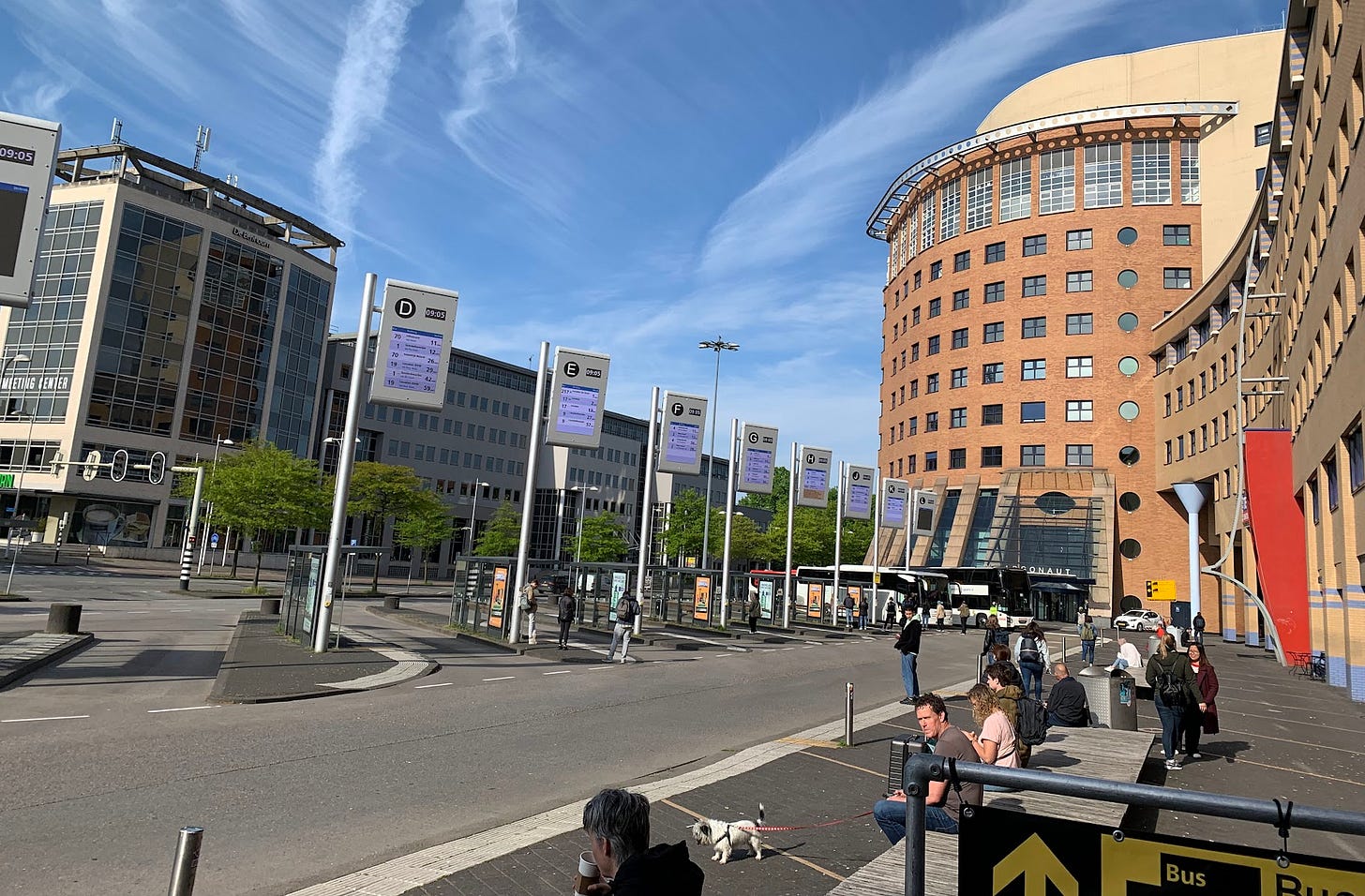 A bus station with many bus platforms visible. Each platform has a large vertical sign with route and times displayed that is visible from a large pedestrian plaza and shopping area perpendicular to all the platforms.