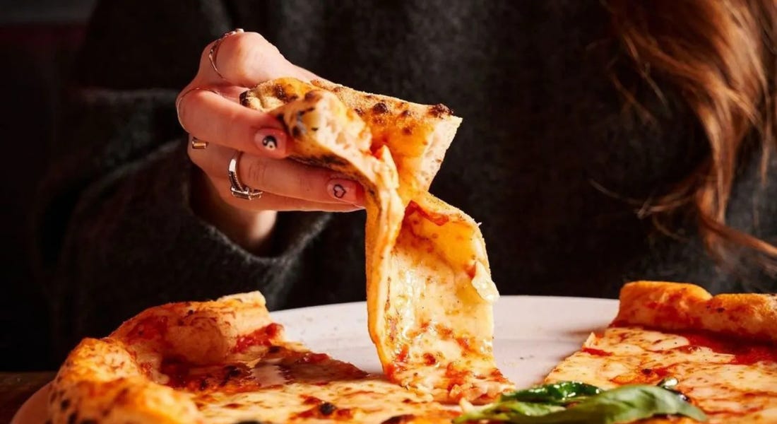 A woman wearing a black jumper holding a pizza slice with a cheese pizza in front of it A woman wearing a black jumper holding a pizza slice with a cheese pizza in front of it