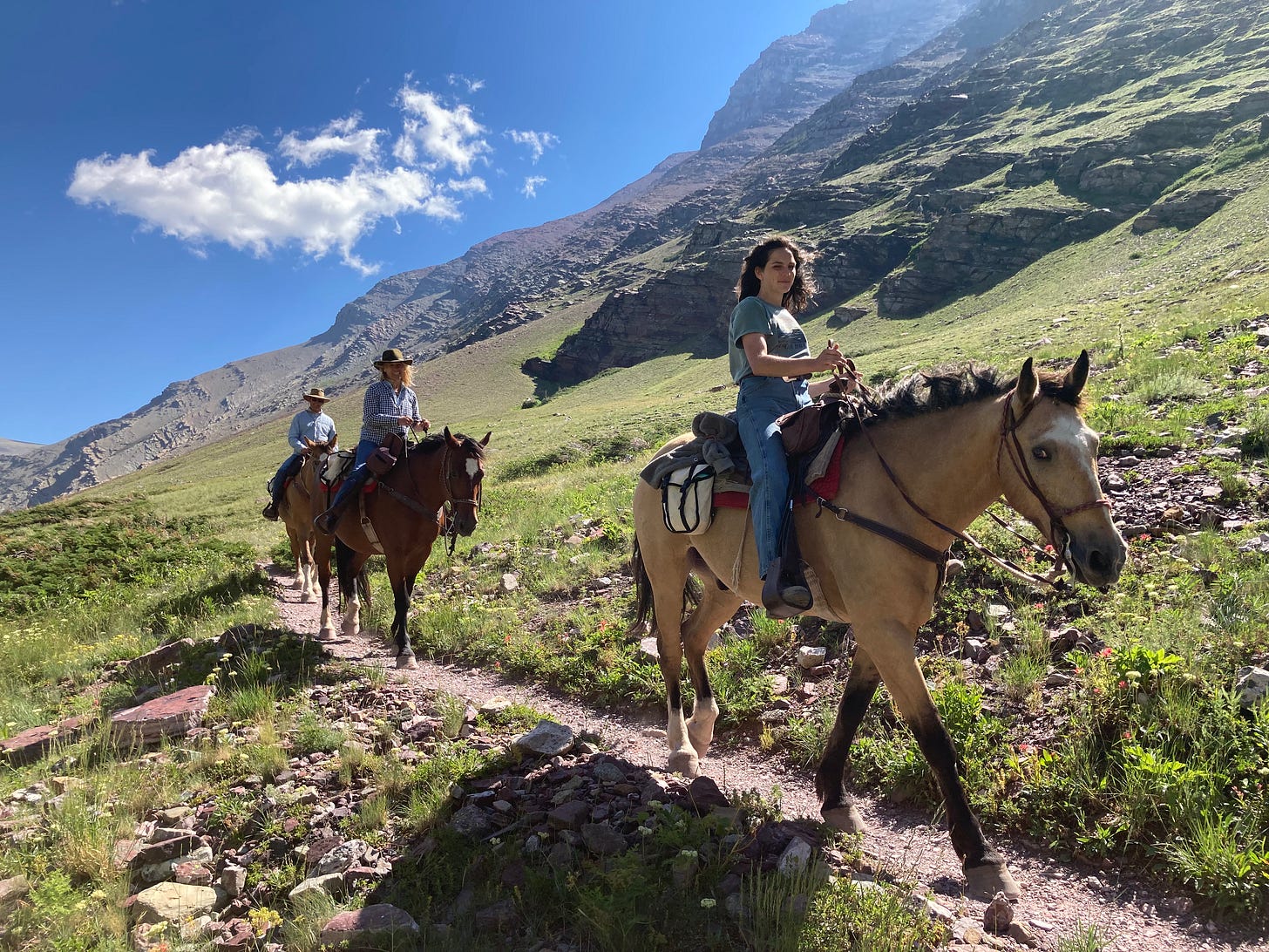 Three horseback riders follow a trail with towering mountain cliffs in the background. Three horseback riders follow a trail with towering mountain cliffs in the background.
