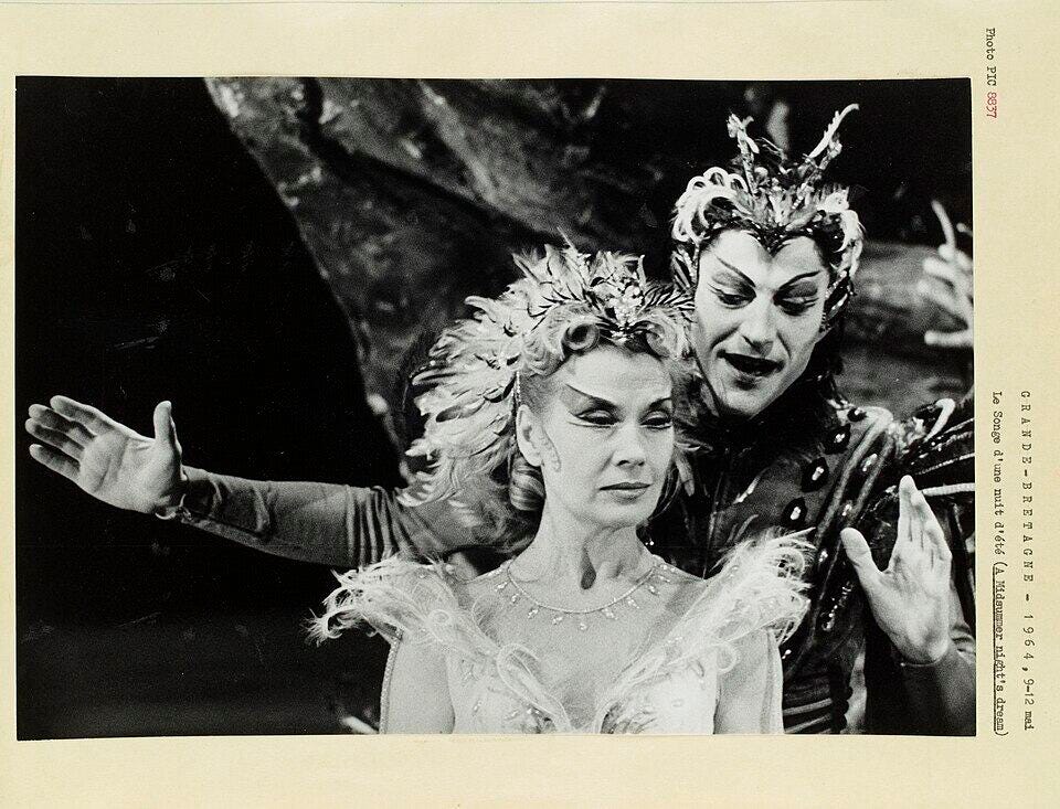 Black and white photograph of a woman in a white dress and elaborate surrealist crown; a dark clad winged figure leans over her shoulder