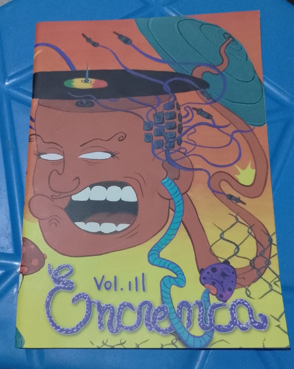 A imagem mostra a capa de uma revista intitulada "Encrenca", especificamente o Volume III. A ilustração central é vibrante, com um estilo artístico que mistura elementos do surrealismo, afrofuturismo e estética cyberpunk. Composição e Cores Cores Predominantes: O fundo é de um tom amarelo-alaranjado quente. A arte utiliza cores contrastantes como azul-turquesa, roxo, verde e tons terrosos. Personagem Central: No centro, há o rosto de uma pessoa negra estilizada, vista de frente e levemente de baixo para cima. O rosto tem uma expressão intensa: a boca está bem aberta, mostrando os dentes, como se estivesse gritando ou cantando. Os olhos são representados apenas por áreas brancas, sem pupilas, o que confere um aspecto místico ou tecnológico. Elementos Detalhados Adereços e Conexões: No topo da cabeça, a pessoa usa o que parece ser um chapéu ou acessório circular preto com detalhes coloridos no centro (verde, amarelo e vermelho). Saindo do lado direito da cabeça e descendo pelo pescoço, há uma série de cabos, fios e conexões que lembram circuitos eletrônicos ou tentáculos tecnológicos, reforçando o tema futurista. Fundo e Texturas: Ao fundo, do lado direito, vê-se uma forma verde que lembra uma folha grande com nervuras, e uma estrutura de rede preta que se sobrepõe a parte da ilustração. Tipografia: Na parte inferior, a palavra "Encrenca" está escrita em uma fonte cursiva, estilizada como se fosse feita de uma corda ou mangueira roxa com pontos azuis. Logo acima da letra "n", está escrito de forma simples: "Vol. III".