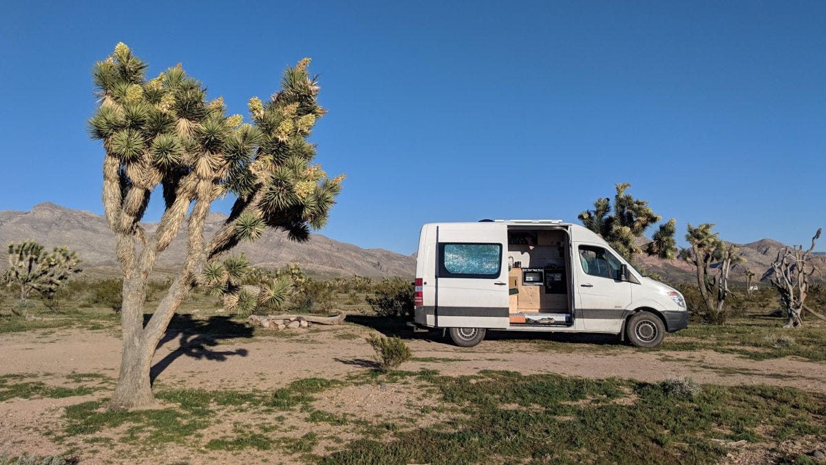 A white van with its side door open is parked on desert ground next to a Joshua tree, embodying the spirit of van life, with mountains in the background under a clear blue sky. A white van with its side door open is parked on desert ground next to a Joshua tree, embodying the spirit of van life, with mountains in the background under a clear blue sky.