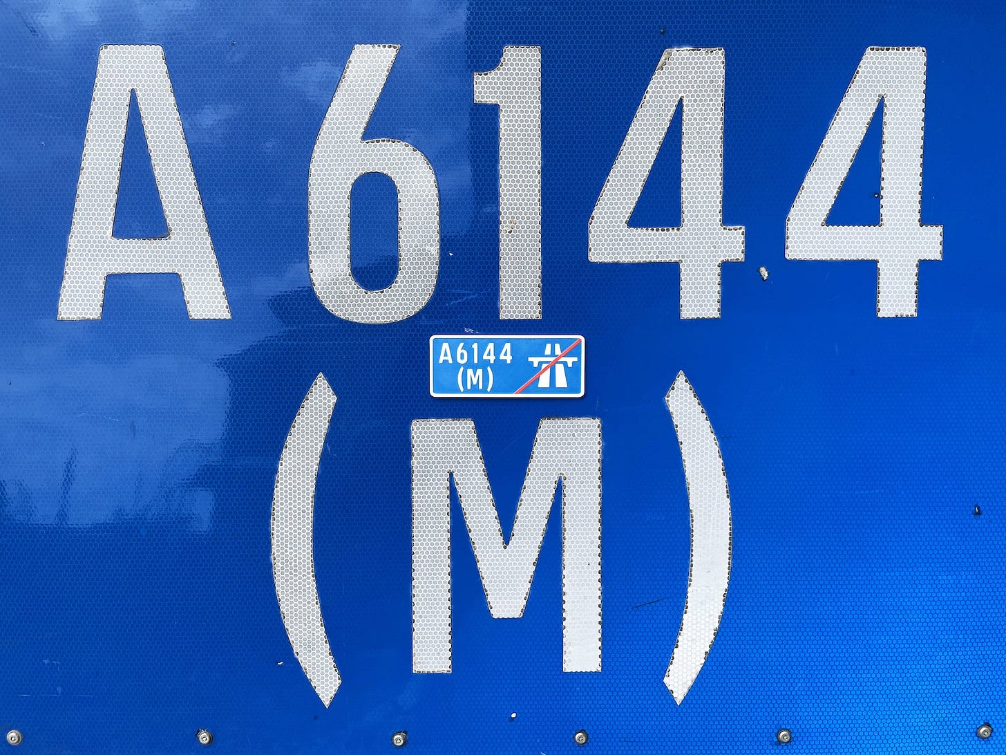 Close-up of a bright blue road sign bearing the text "A6144(M)". Between the digits and the "(M)" is placed a small fridge magnet, which is a replica of the sign. Close-up of a bright blue road sign bearing the text "A6144(M)". Between the digits and the "(M)" is placed a small fridge magnet, which is a replica of the sign.