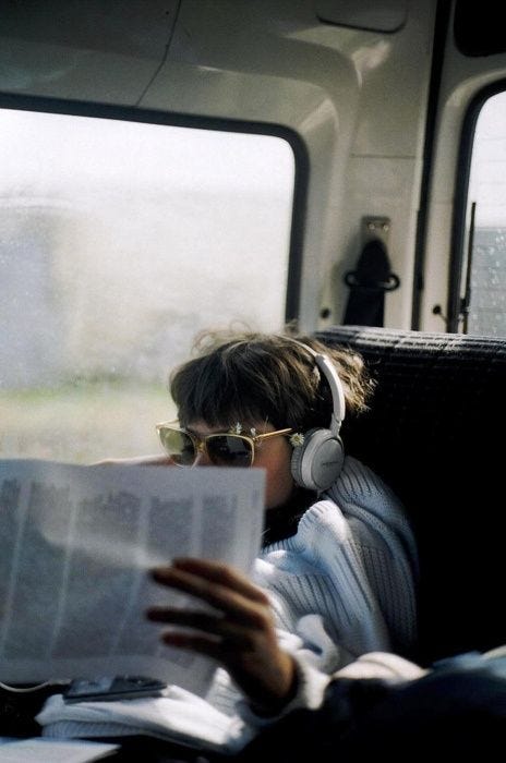 A man wearing headphones and sunglasses reads a newspaper while traveling on a van.