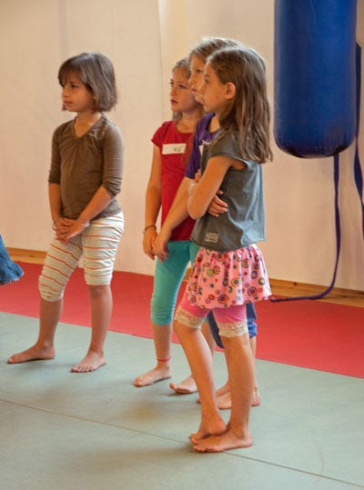 Four small girls are in a gym and looking intently at a situation out of frame.   