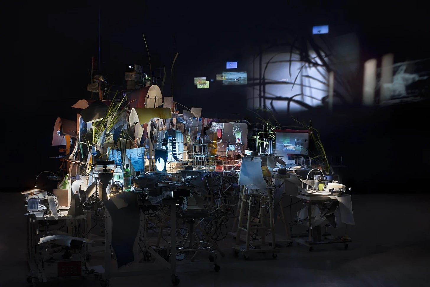 A view of Sarah Sze's installation Timekeeper (2016). Emerging from the background of a dark room is a kind of deskscape lit by projected images on many varying surfaces. On the wall you can see a projection of grasses. The desk is covered by a jumble of objects including plants, fans, clocks, and lights