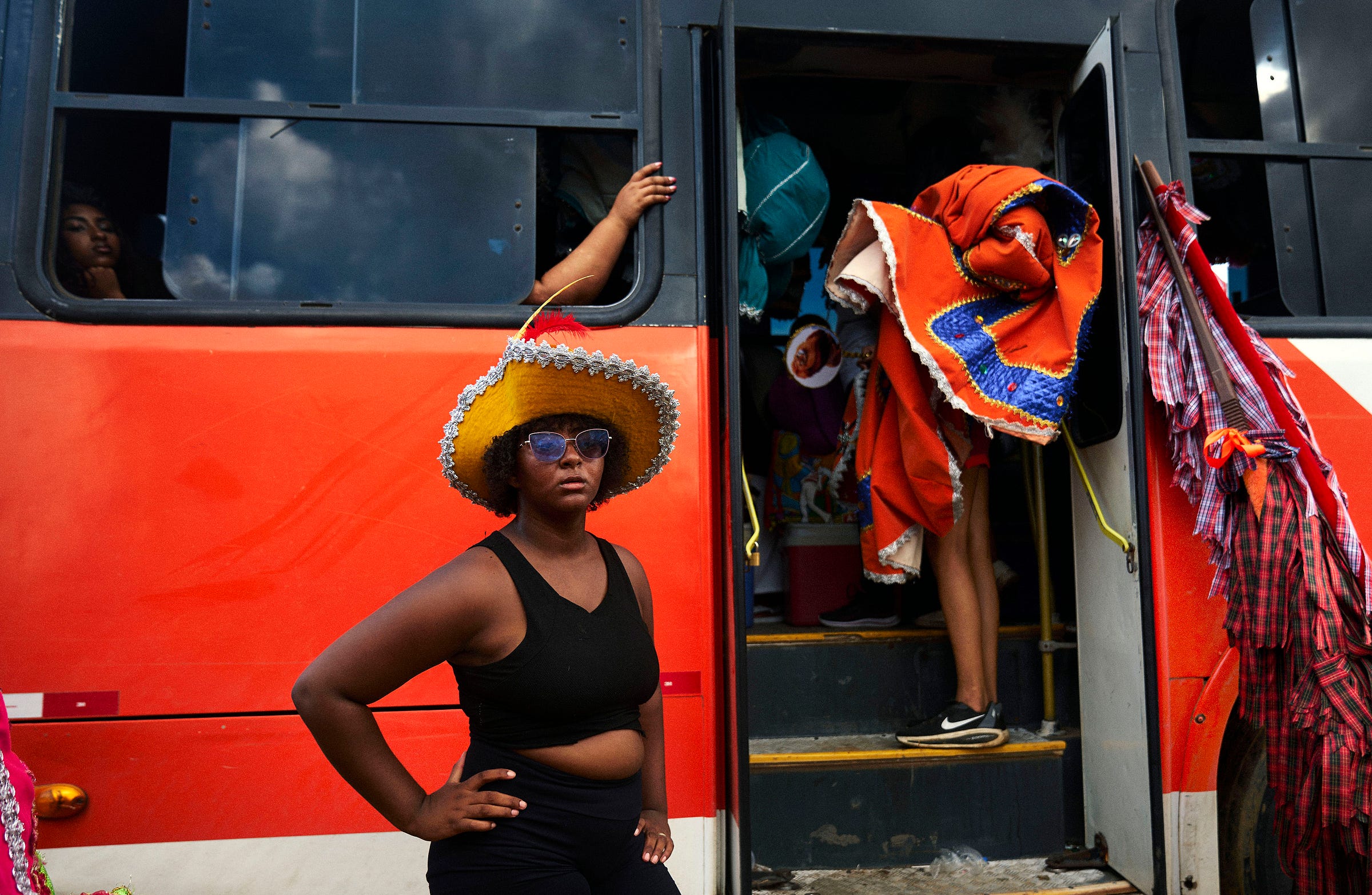 A woman in a yellow hat and sunglasses stands in front of a red bus. Another person draped in a colorful blanket boards the bus, while others are visible inside through the windows.