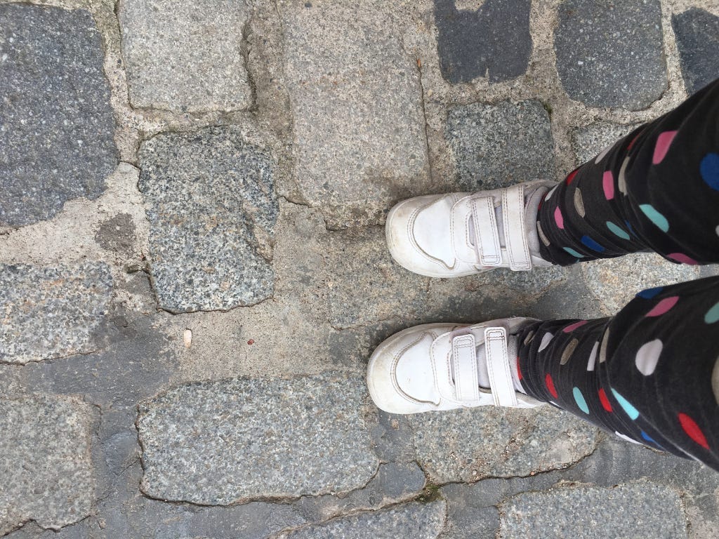 a toddler with her shoes on the wrong feet standing on a cobblestone street