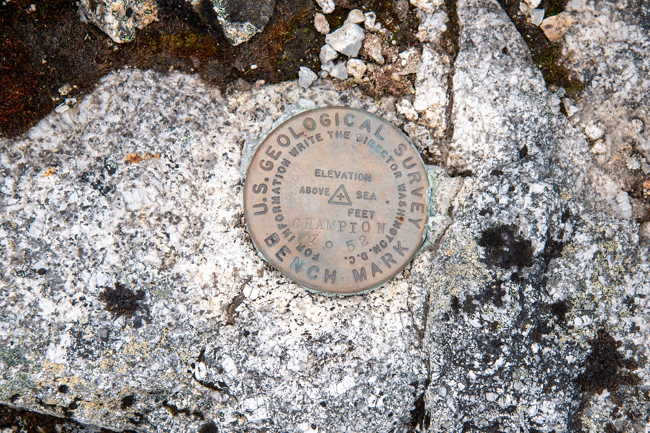 A round bronze USGS benchmark disk set into pale, lichen-blotched granite, stamped U.S. Geological Survey Bench Mark with a triangle at its center and the name Champion and the year 1952.