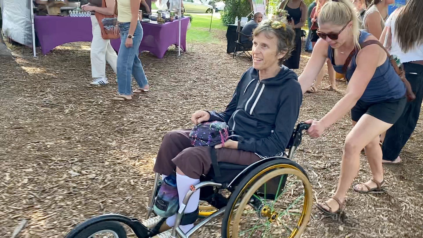 A woman with short brown hair wearing brown pants and a navy blue sweatshirt is pushed in her wheelchair by a woman with long blond hair in a ponytail wearing black shorts, navy tank top, sunglasses and sandals at a farmers market.