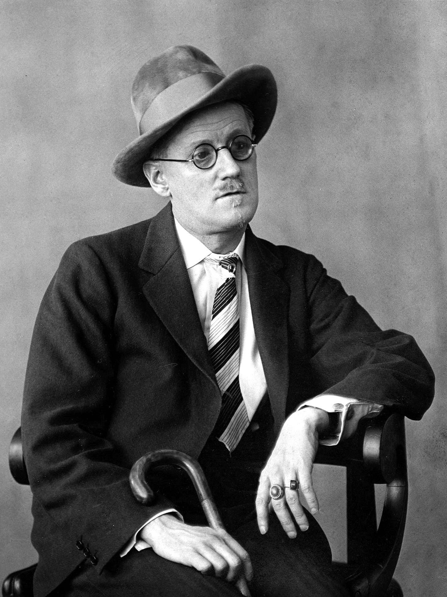 A man in a suit, tie, and hat sits on a chair holding a cane, wearing round glasses and looking slightly to the side.