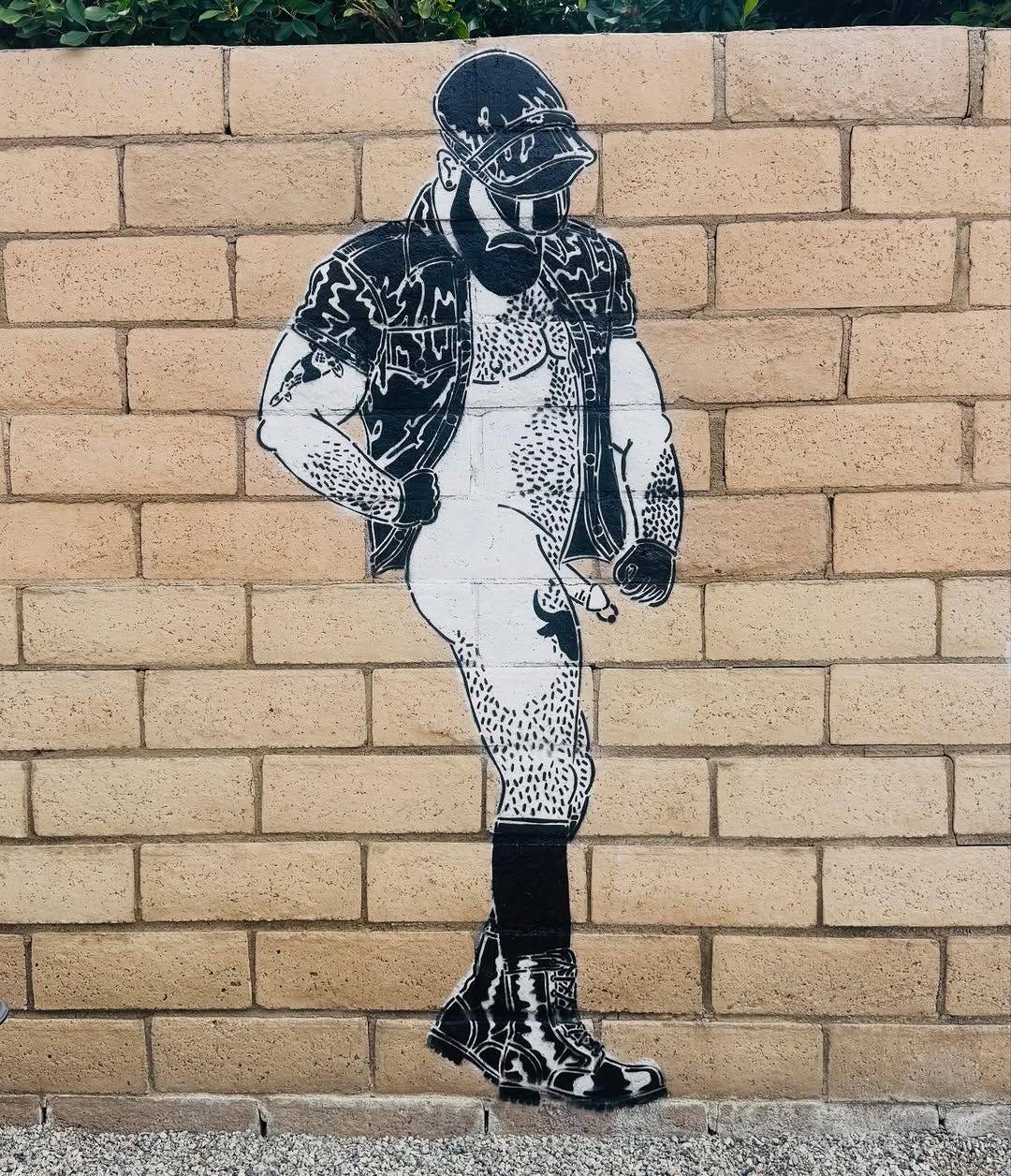 Street art of a person with a black cap, beard, sunglasses and leather gear painted on a brick wall. The bold lines and stance convey a confident, edgy tone. The man is mostly nude, with his dick sticking out. He has hairy arms, legs, and torso, and he is wearing black knee-high socks and boots, with gloves on each hand. He is looking down at his groin. His leather jacket is open, revealing his bare chest. Street art of a person with a black cap, beard, sunglasses and leather gear painted on a brick wall. The bold lines and stance convey a confident, edgy tone. The man is mostly nude, with his dick sticking out. He has hairy arms, legs, and torso, and he is wearing black knee-high socks and boots, with gloves on each hand. He is looking down at his groin. His leather jacket is open, revealing his bare chest.