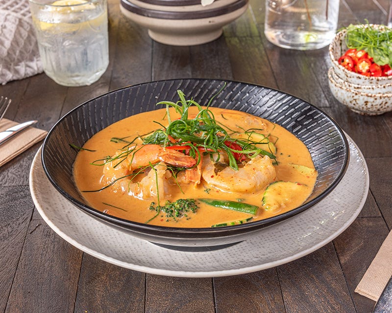 A black bowl atop a white plate, filled with yellow-orange curried prawns, garnished with green A black bowl atop a white plate, filled with yellow-orange curried prawns, garnished with green
