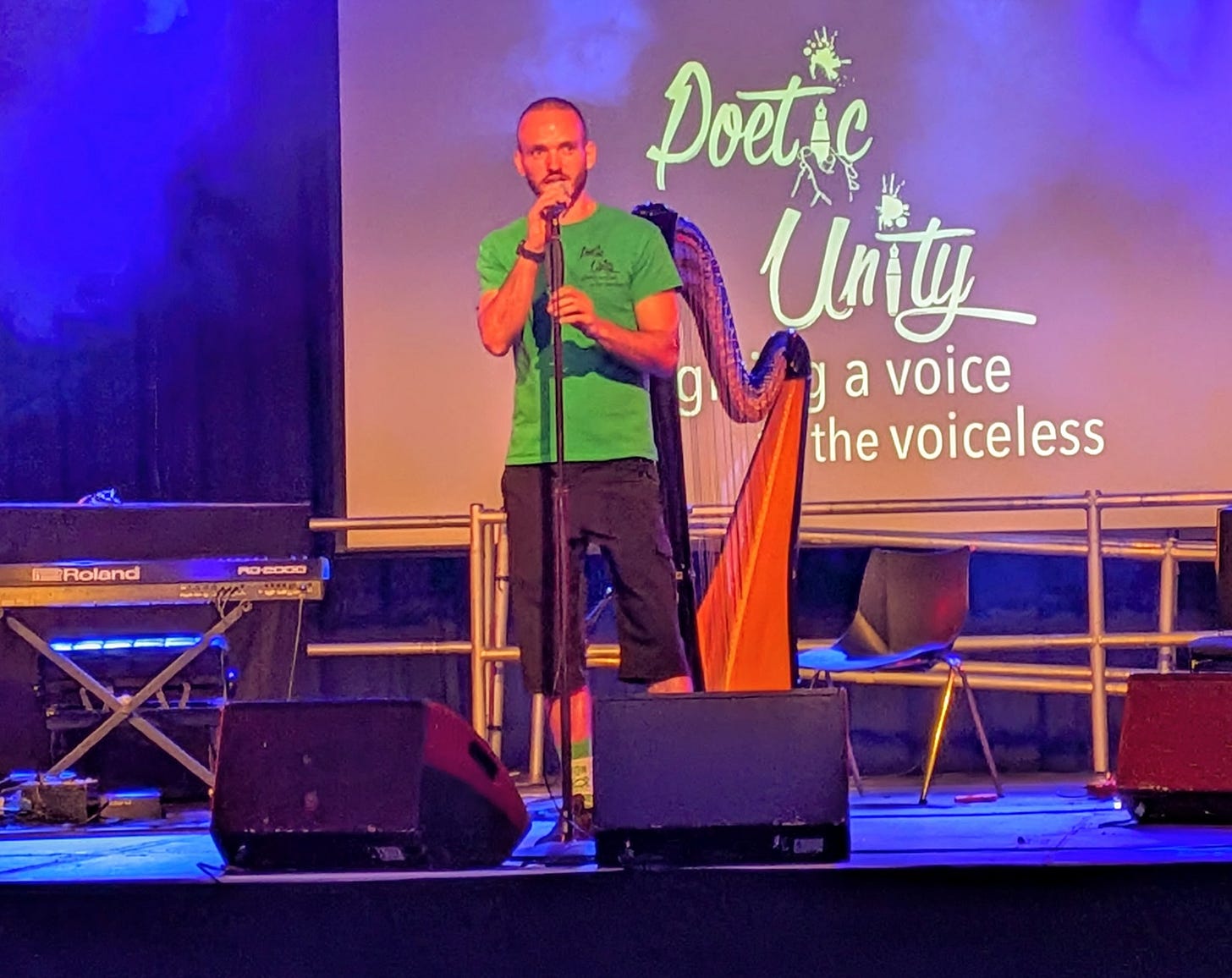 RagCV from Poetic Unity on a stage at the Southbank Centre. He is wearing a green t-shirt and there is a harp on the stage behind him. Also behind him is a banner that says 'Poetic Unity, giving a voice to the voiceless'