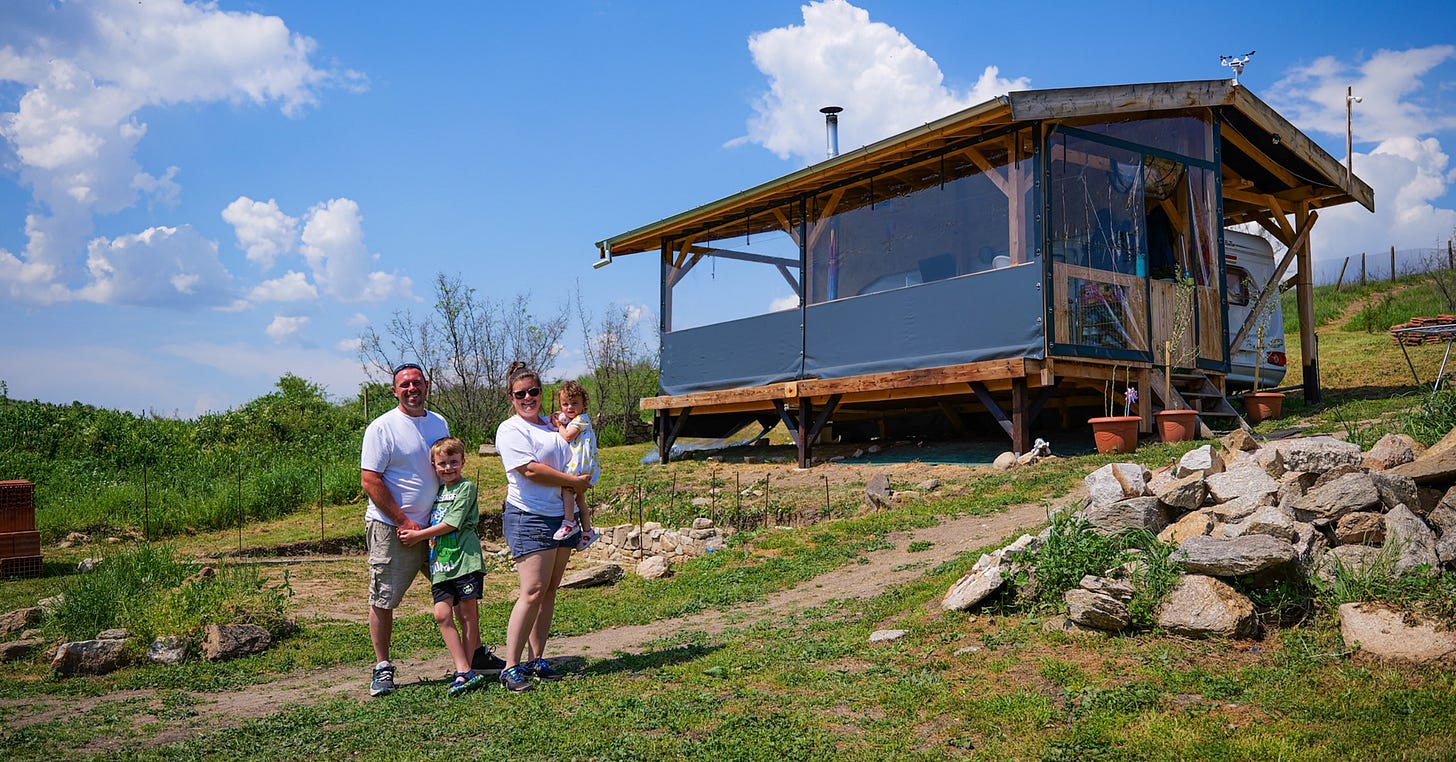 A family stands on a grassy area, with a wooden house featuring large windows in the background under a blue sky with fluffy clouds.