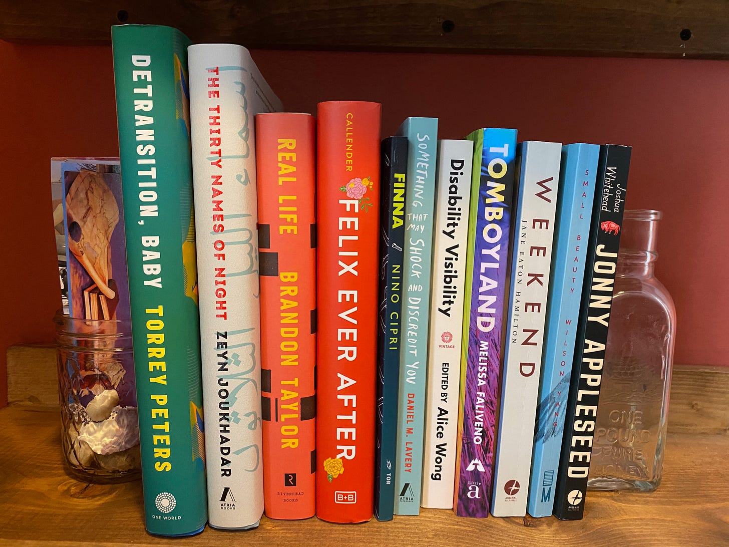 A row of books on a wooden shelf. The books are a variety of shapes and colors, and are bookended by a glass jar full of bookmarks on one end and an empty glass bottle on the other.