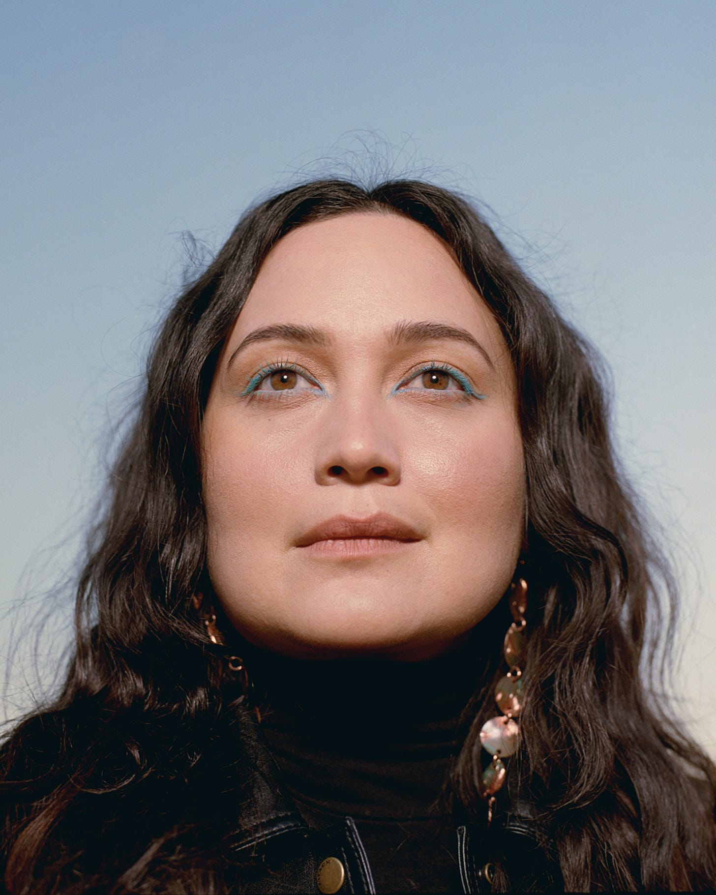 Portrait of Lily Gladstone looking up beyond the camera. Gladstone is wearing a black leather jacket a black turtleneck...