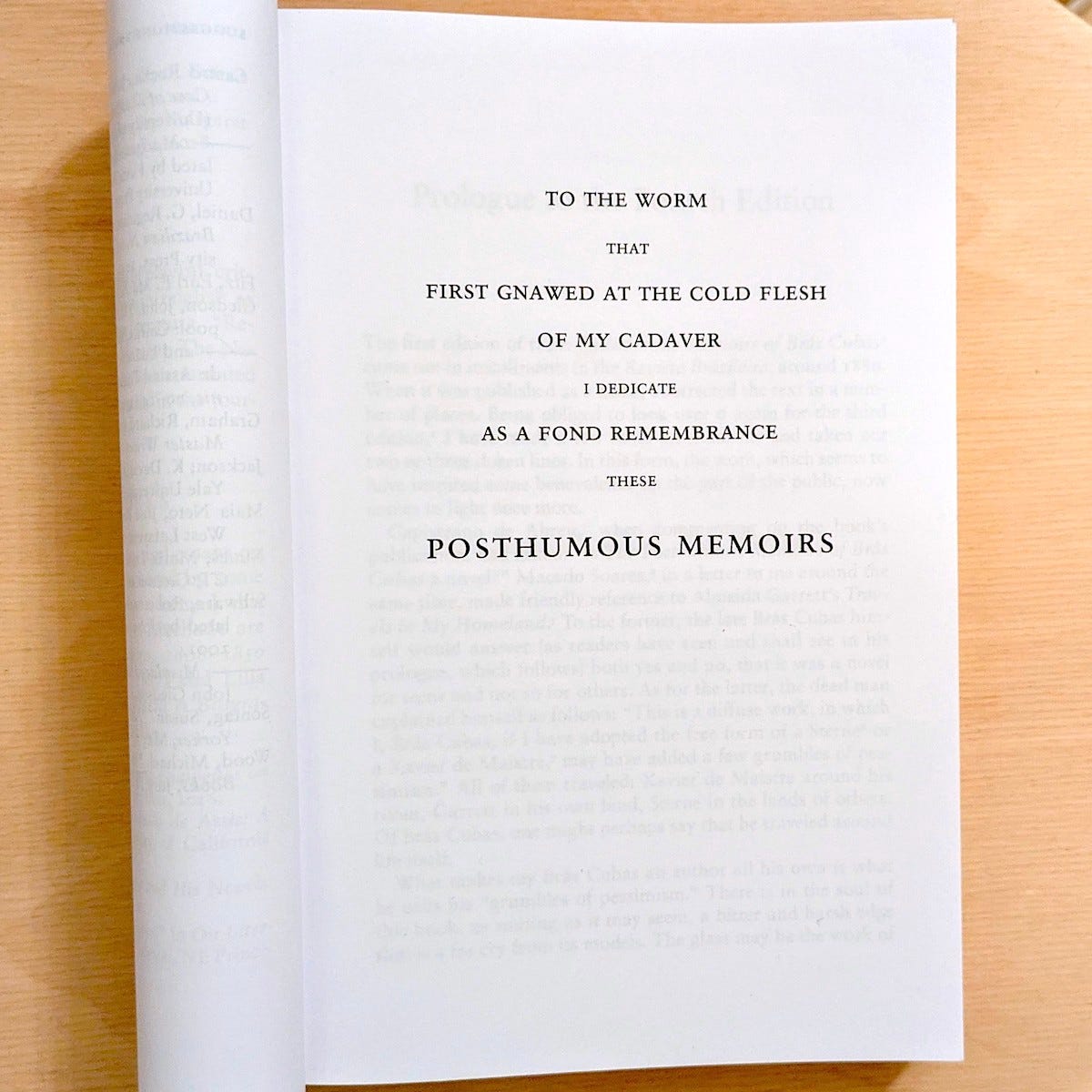 Photo of an open book, on a wooden surface. On the visible page of the book, in all caps: To the worm // that // first gnawed at the cold flesh // of my cadaver // I dedicate // as a fond remembrance // these // posthumous memoirs.