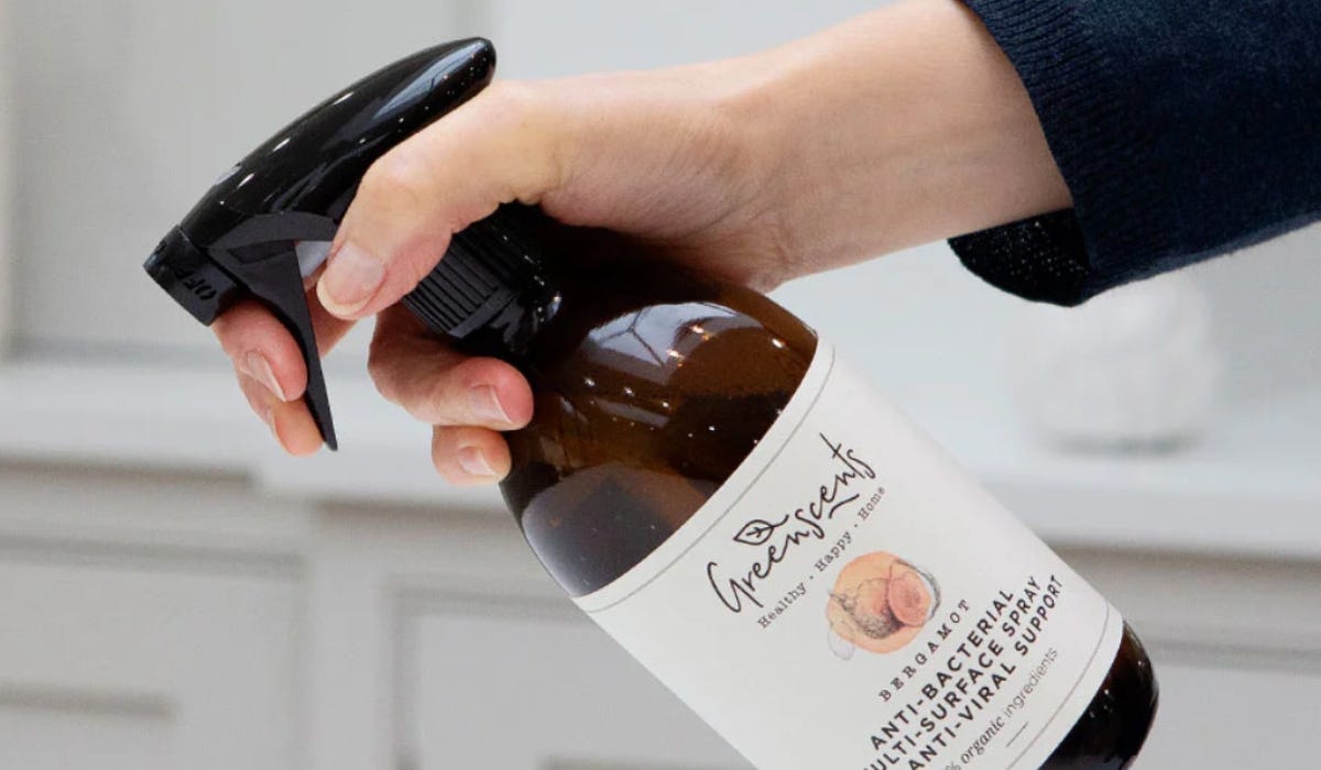 A spray bottle of organic anti-bacterial spray by Greenscents, being held by someone wearing a black long-sleeved top against white cupboards.