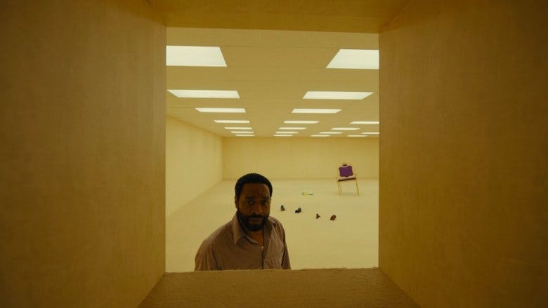A still from the film "Backrooms," in which Chiwetel Ejiofor stands in a square-sized doorway with a fluorescent-lit room behind him. The room has a throne-like chair and some shoes sticking out of the floor. 