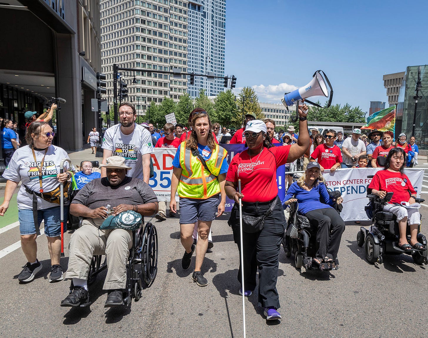 A row of seven marchers lead a group of 1400 people out of Boston City Hall plaza. Among them are wheelchair users, blind walkers, and others: people of all ages, races, and disabilities.