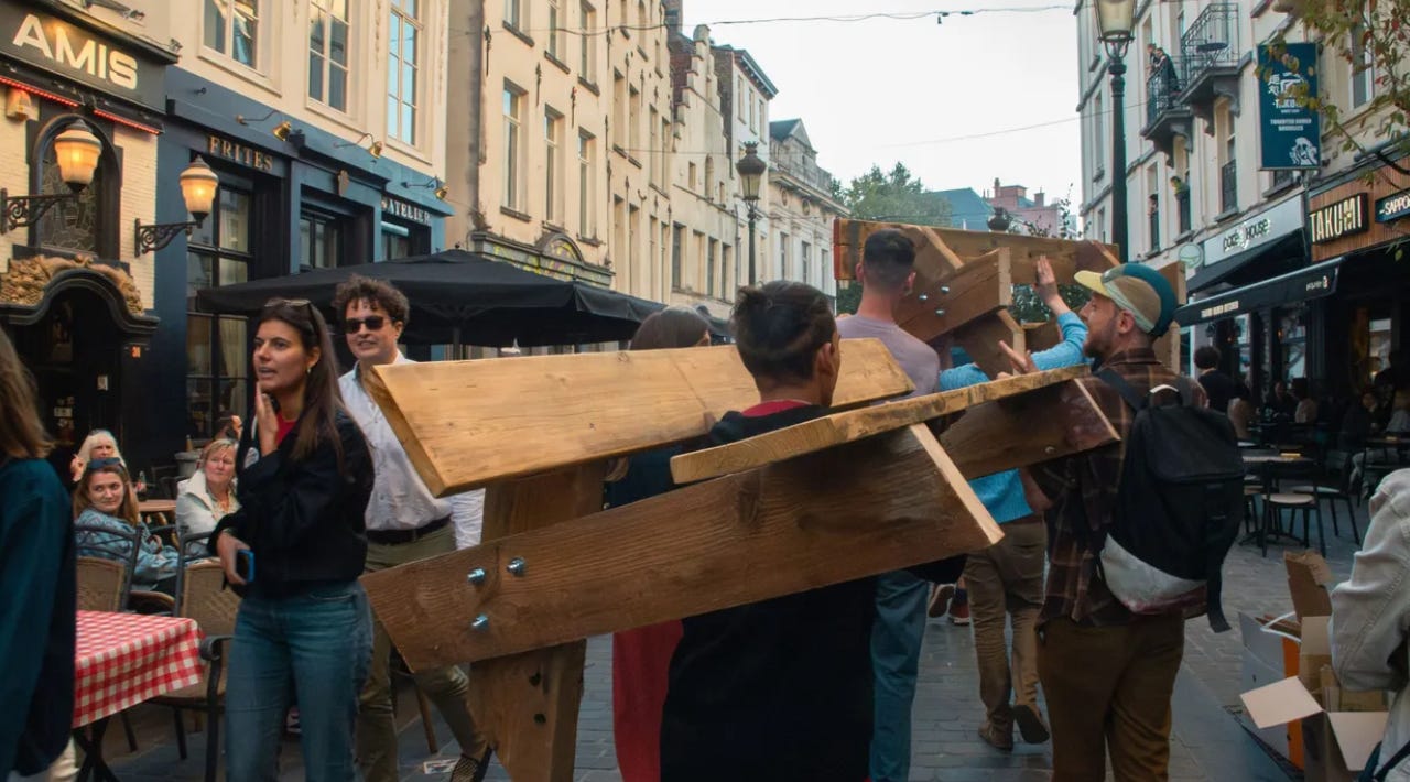 A group of people holding wooden benches and walking through a busy city centre in brussels past some bars. A group of people holding wooden benches and walking through a busy city centre in brussels past some bars.