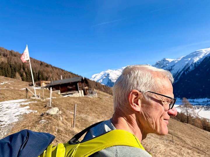 Left image: An elderly woman wearing sunglass clips, a thin down vest and red backpack holds out a hand and points to distant mountain peaks—specifically, the Matterhorn, barely visible in haze. Around her are winter-bare larch trees. The right image, the profile of a white-haired man in glasses with a chartreuse backpack. He views a snow-covered alpine valley floor. Behind him are snow-covered mountains, a winter-brown alpine slope and forest as well as an alpine chalet and a tattered Canton Valais flag, wind-whipped.