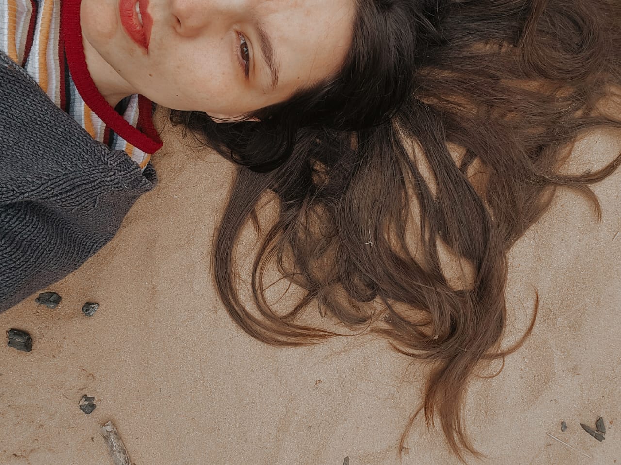 A woman lies on sand, only half her face visible. Her hair spills like smoke, her lips part slightly - caught between silence and eruption. She's unarmored, unposed, undone. A visual echo of the piece: raw, holy, chaotic, whole.