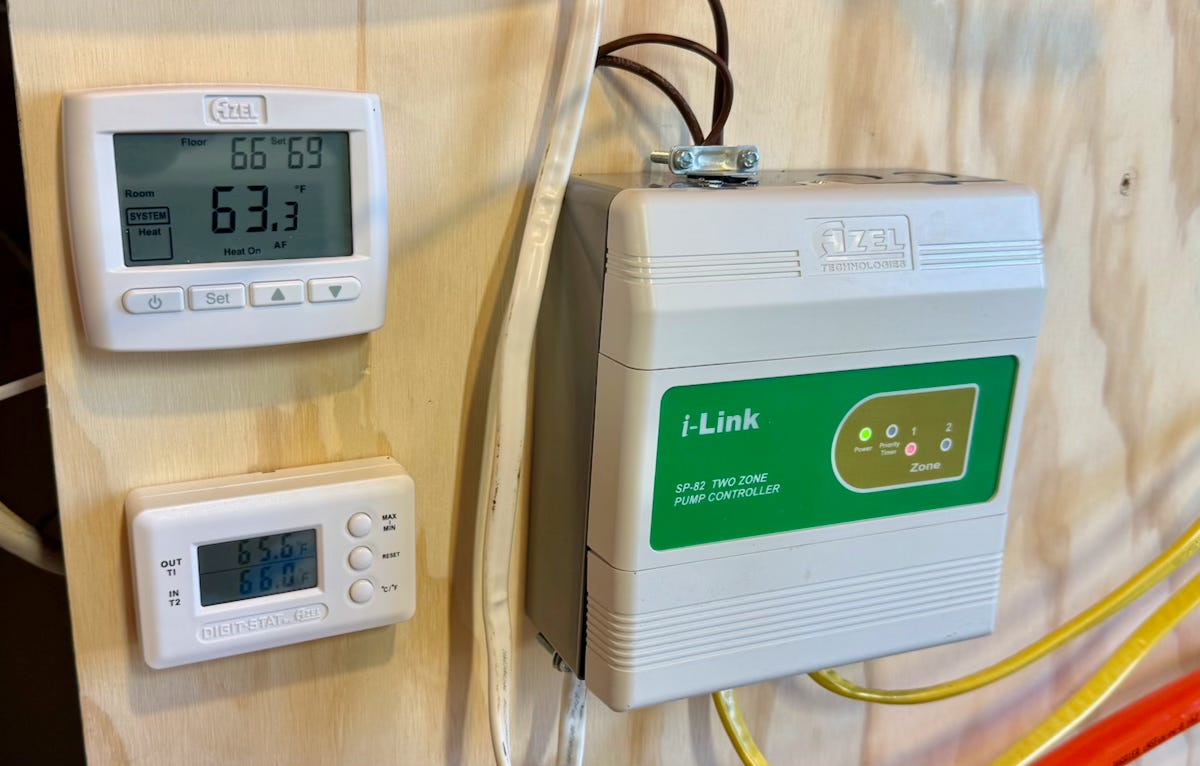 A thermostat, temperature display, and two-zone pump controller are mounted on a plywood wall, showing active readings and controls for a hydronic radiant heating system.