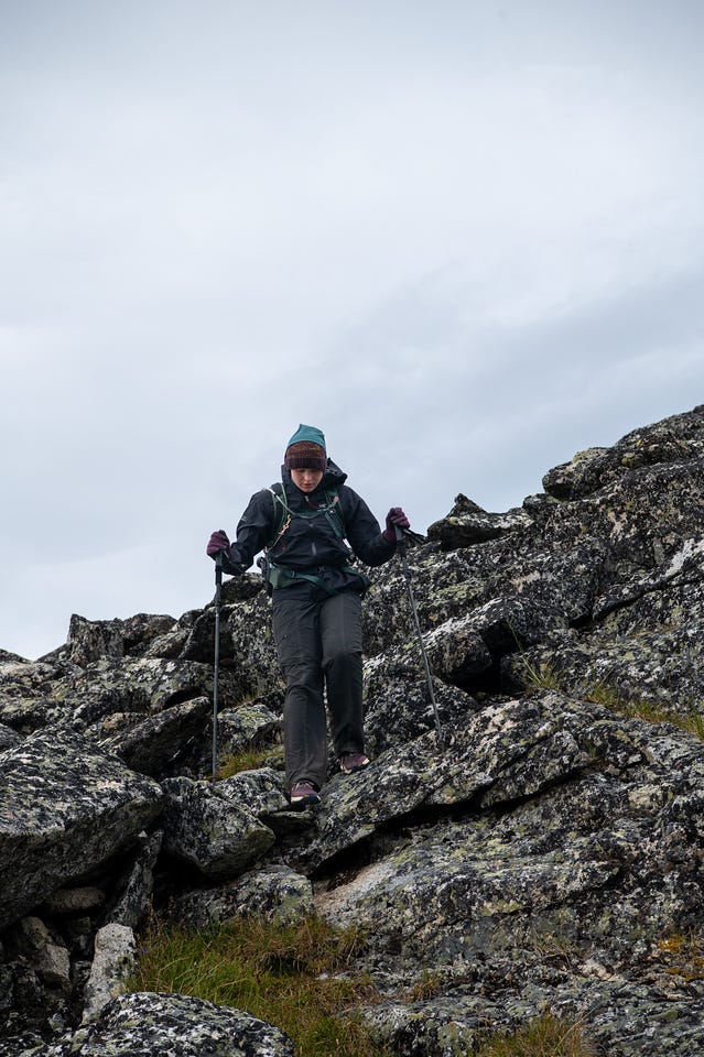A hiker with poles and a dark shell picks down a steep jumble of pale, lichen-mottled talus blocks toward the camera, head down watching her footing, under a flat grey sky.
