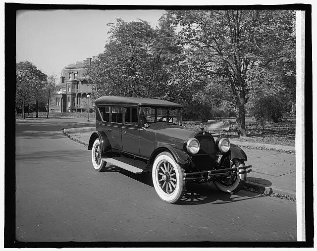 a black and white photo of a 1920s car on the road infront of a large building a black and white photo of a 1920s car on the road infront of a large building
